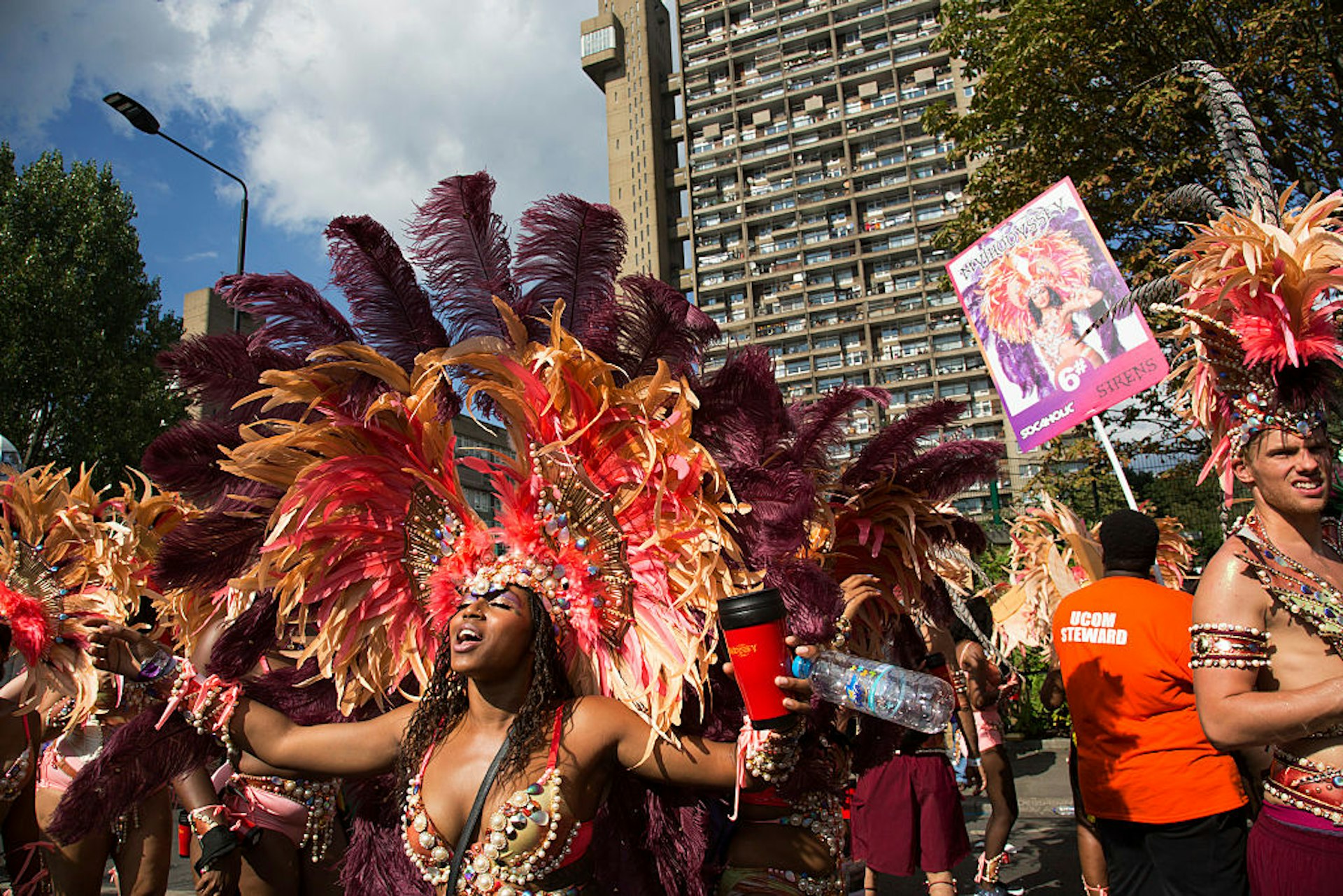 Parade dancers in feather costumes stop in the street at the base of Trellick Tower